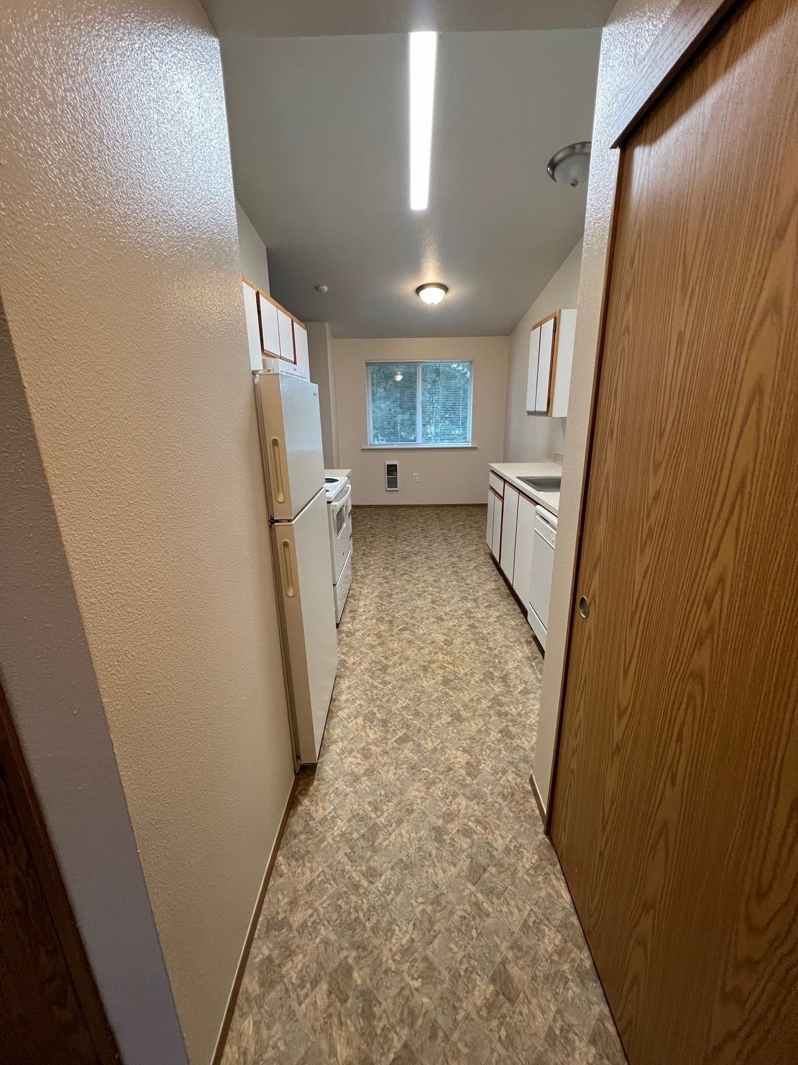 Narrow beige kitchen hallway with white cabinets, wood door, fluorescent light, and a window at the end