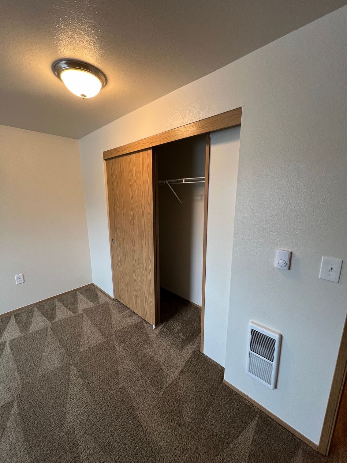 Small carpeted bedroom with open closet, white walls, ceiling light, and wall vent.