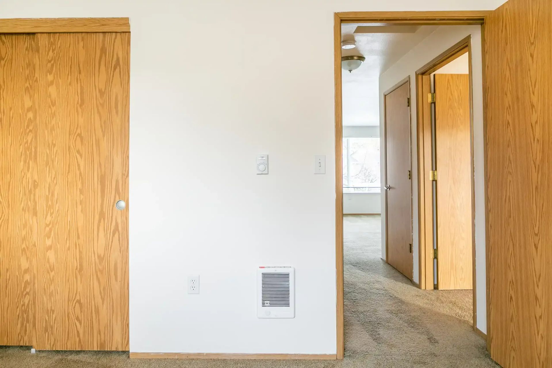Empty room with white walls, wood doors, and a doorway to a carpeted hallway.