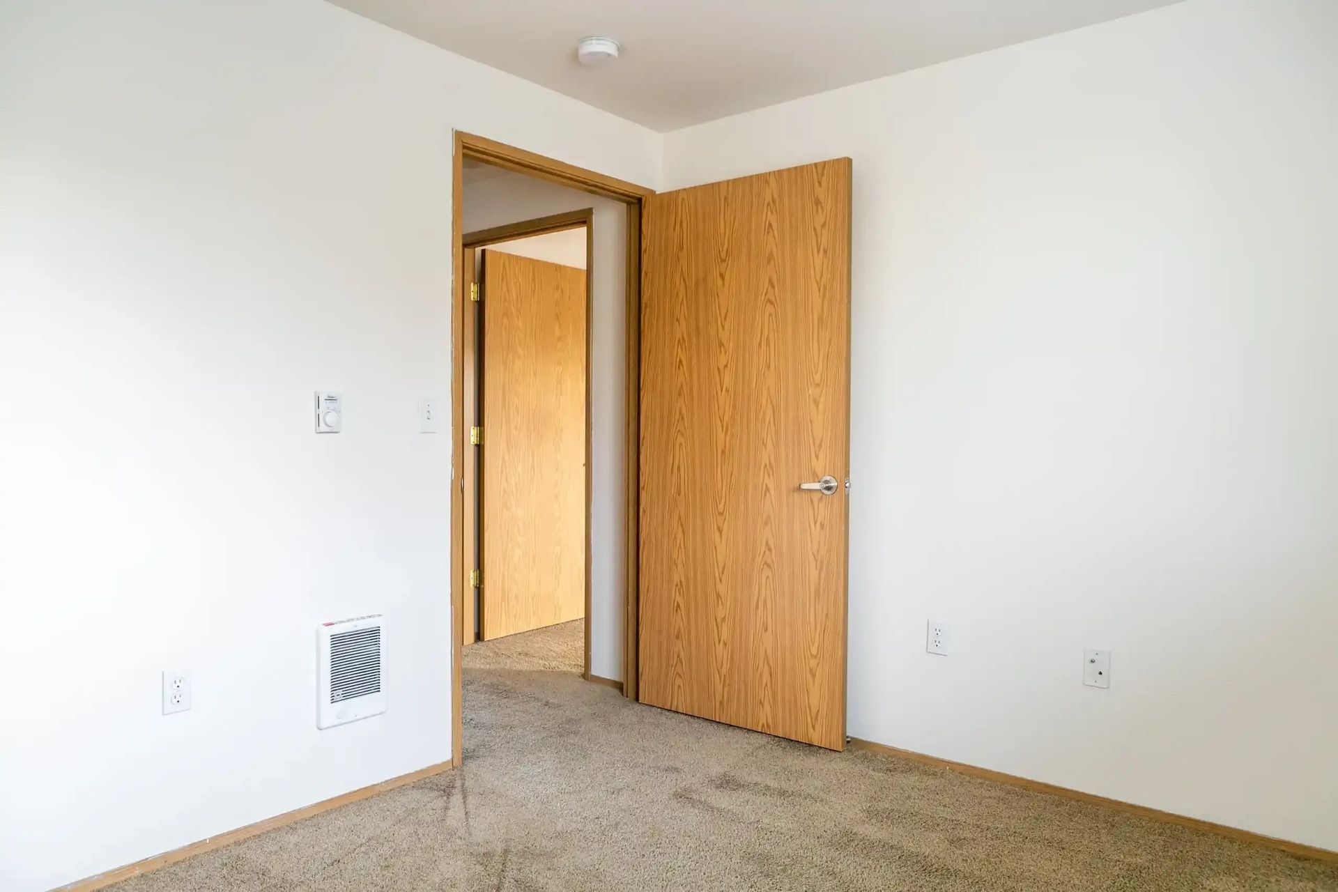 Empty carpeted room with white walls and an open wooden door leading to a hallway.