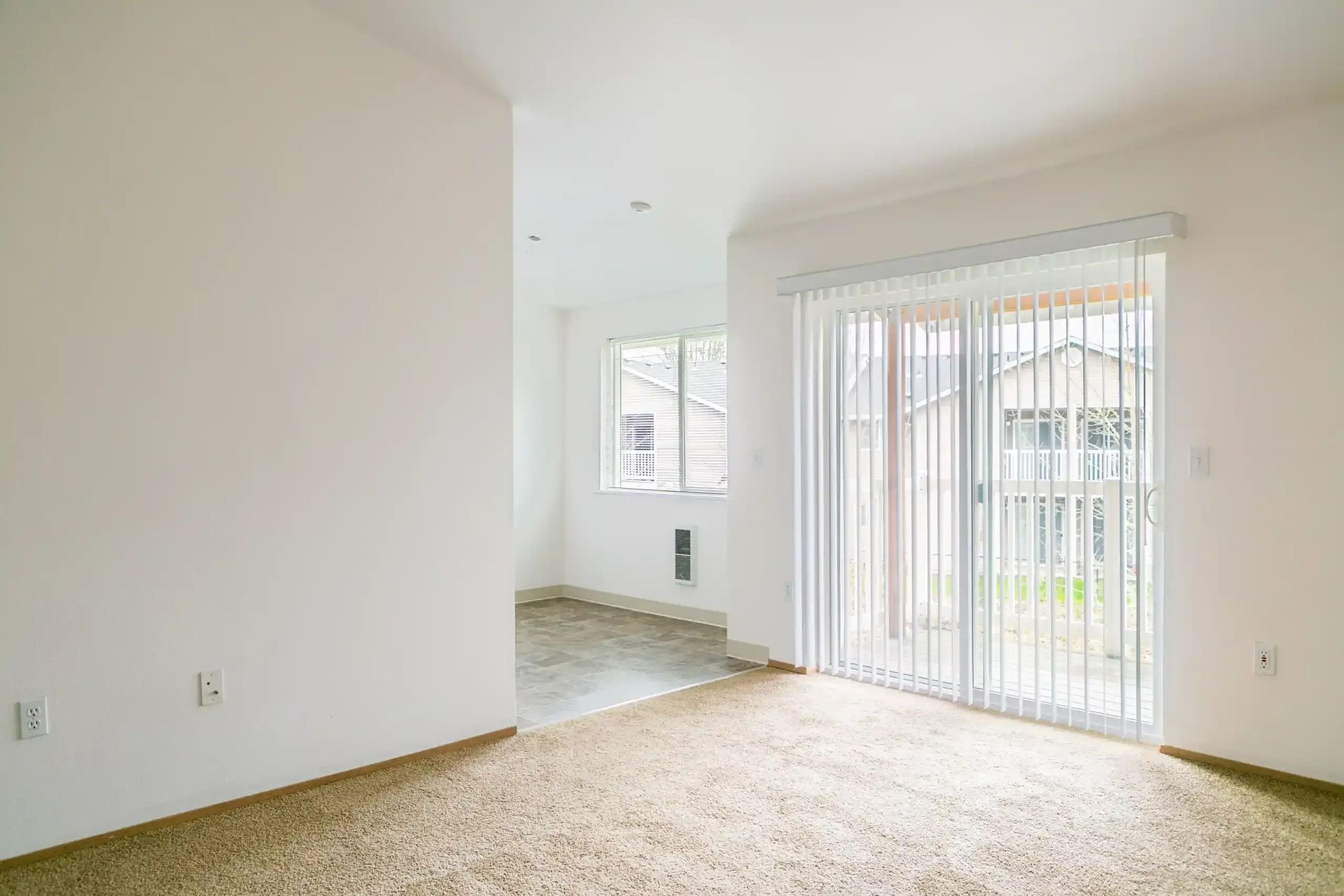 Empty bright living room with beige carpet, white walls, and sliding glass door to a patio