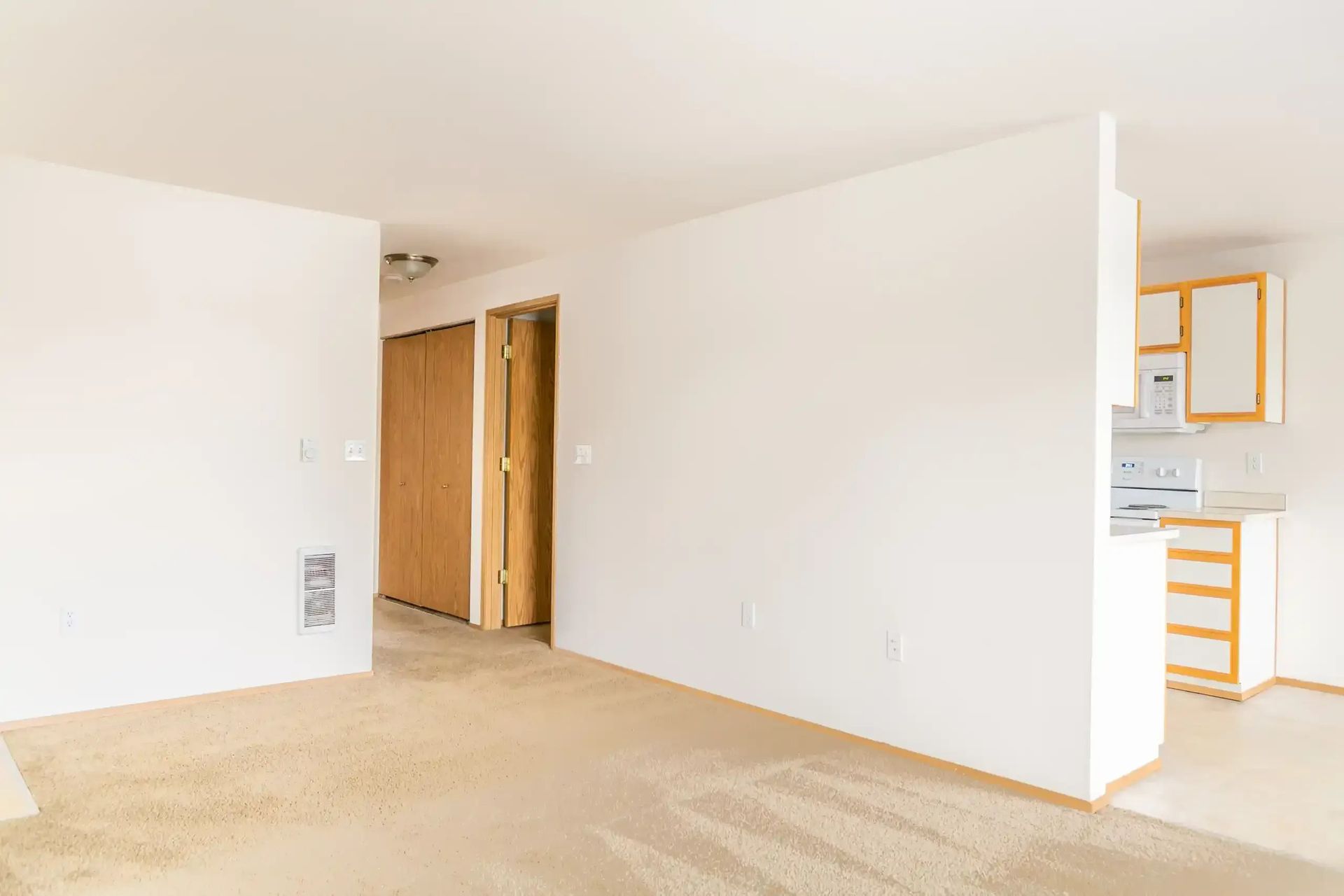 Empty white interior room with beige carpet, wood closet doors, and a small kitchen visible on the right