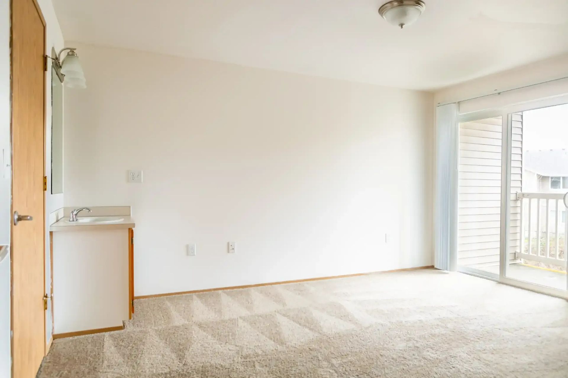 Empty beige living room with carpet, white walls, and sliding glass door to a balcony