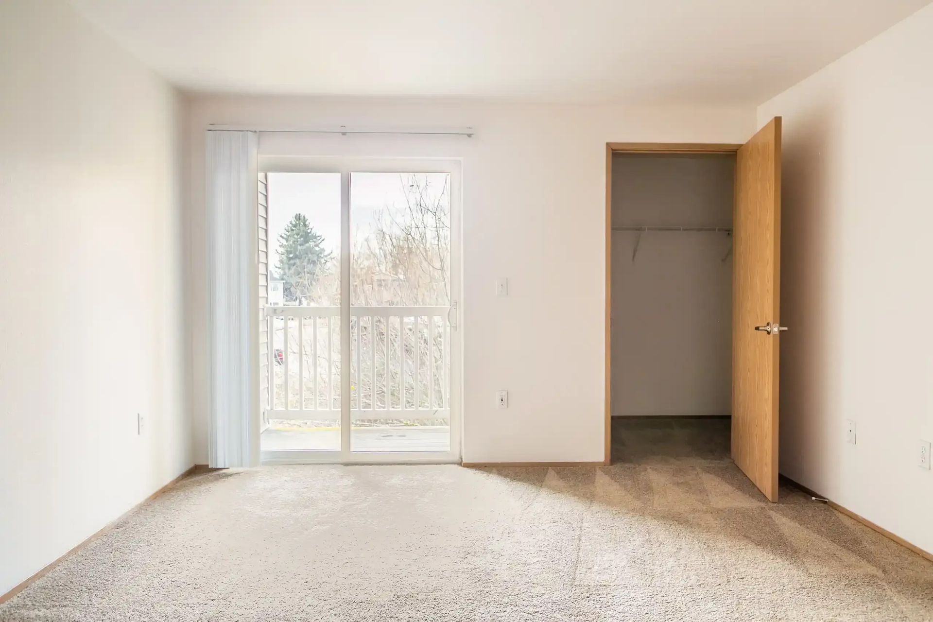 Empty beige bedroom with open closet and sliding glass door to a balcony.