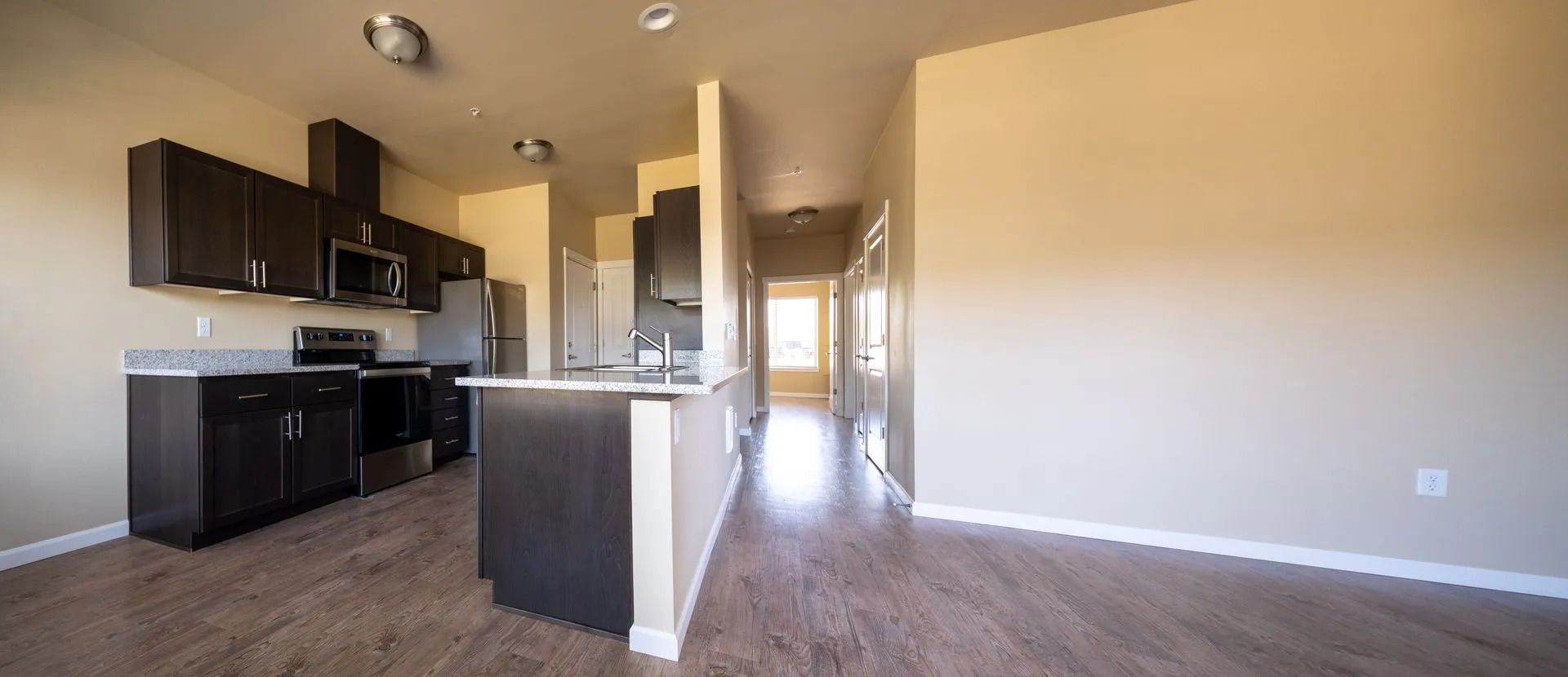 Empty modern apartment with dark kitchen cabinets, island, and beige carpeted living area