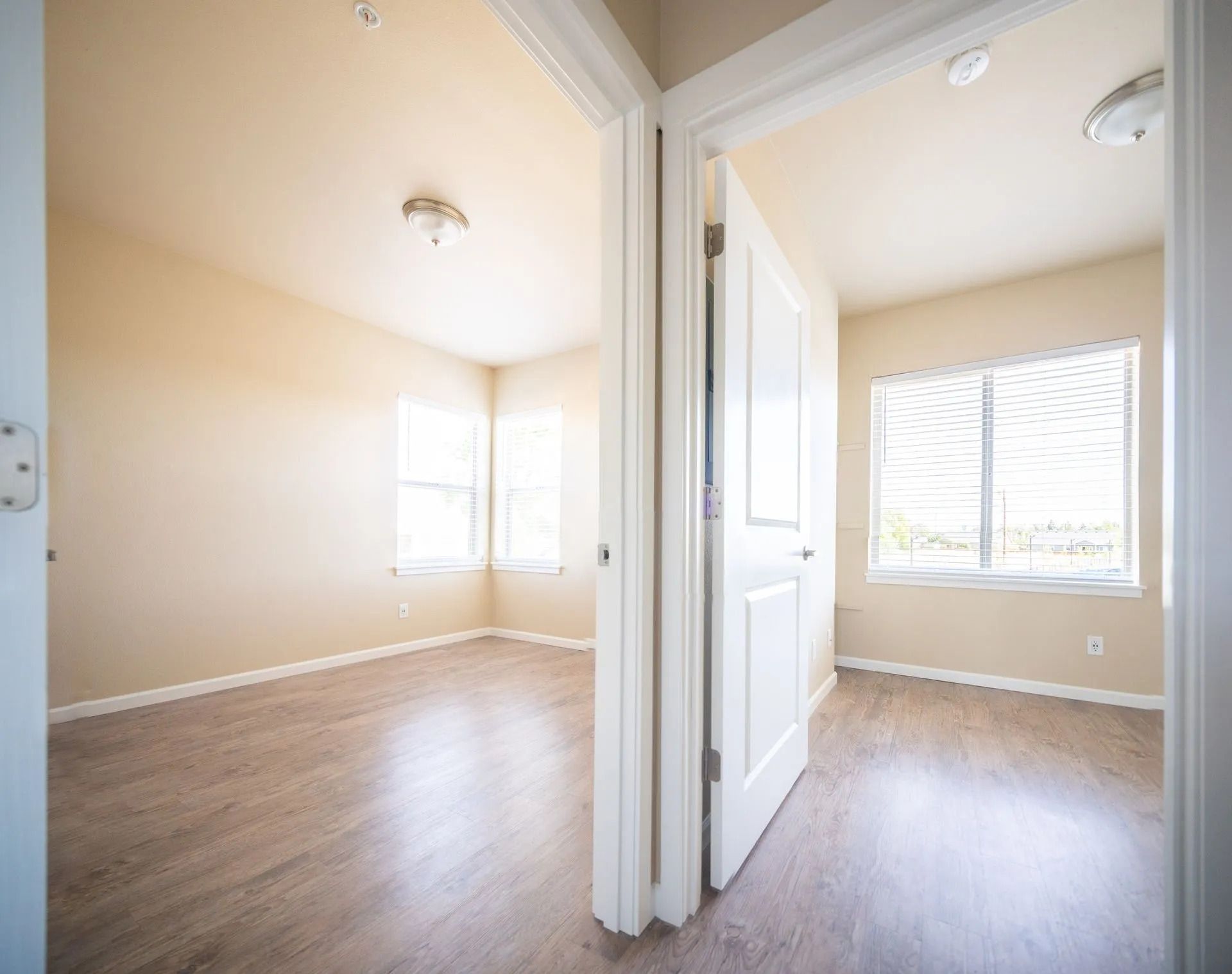 Empty beige apartment rooms with wood floors, white trim, and bright windows through open doorway