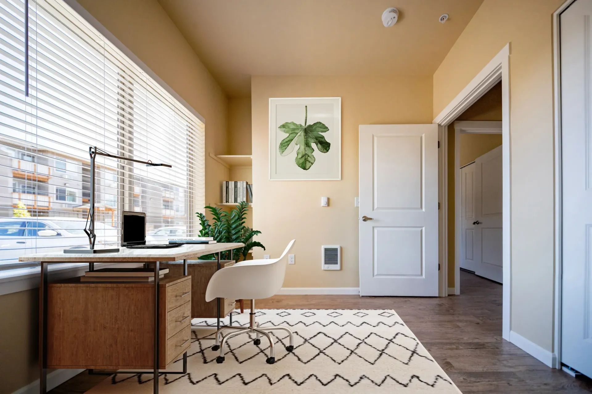 Bright home office with a wooden desk, white chair, plant art, and large window blinds