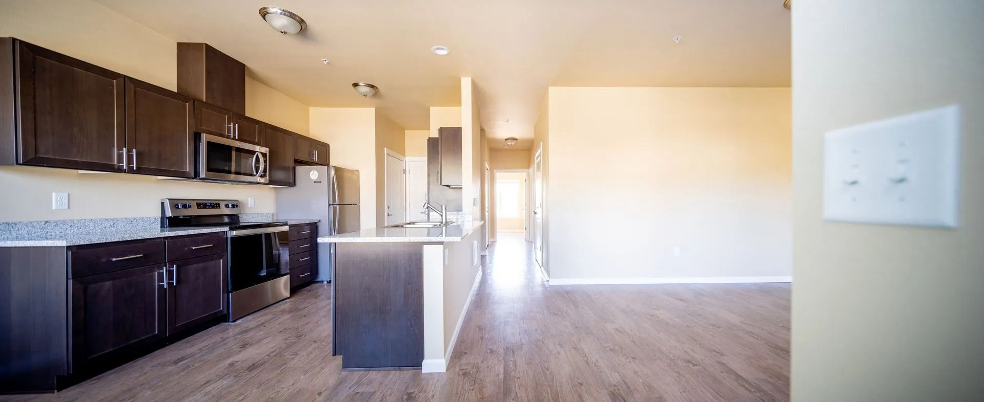 Empty modern apartment kitchen and living area with dark cabinets, island, and wood floors