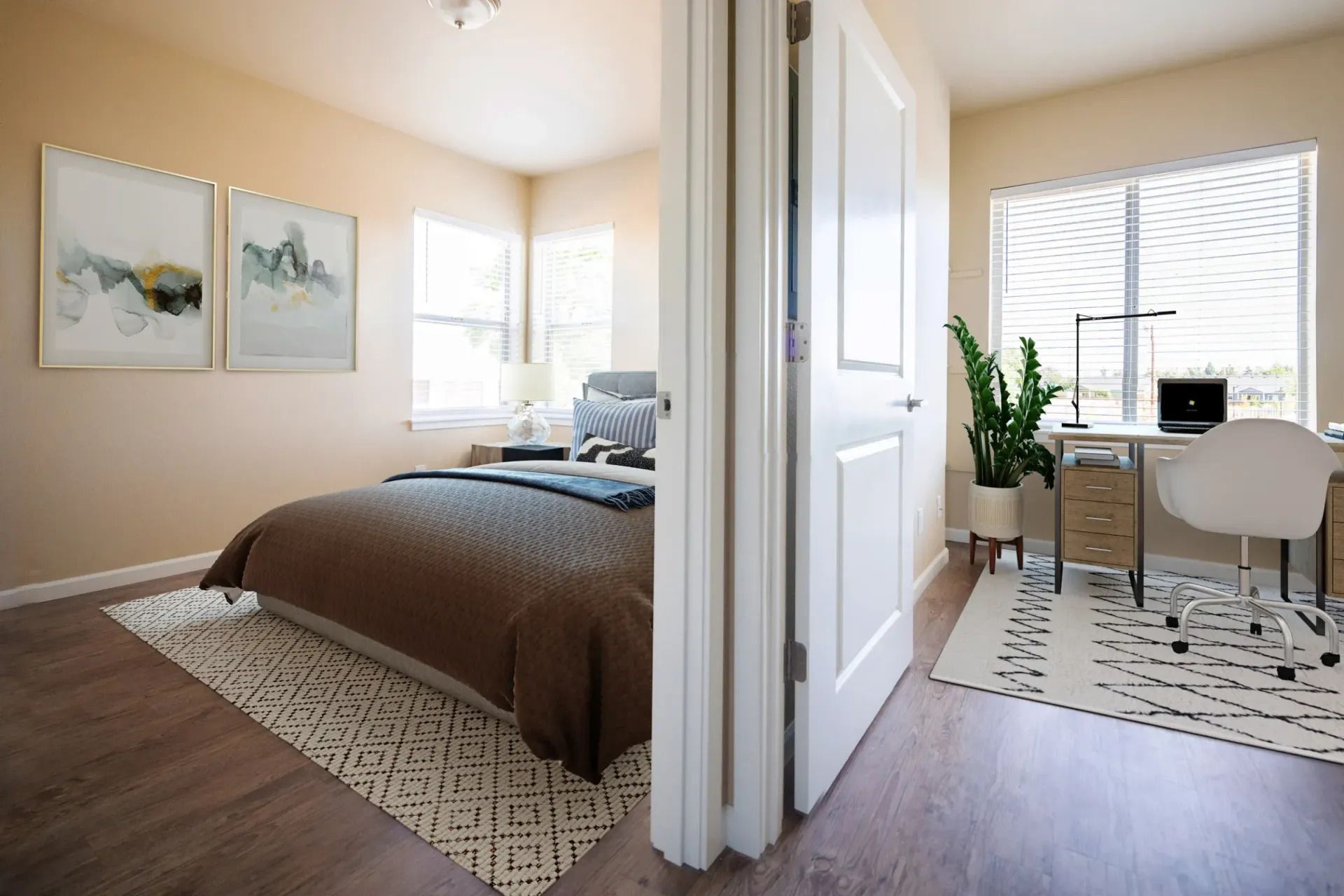 Bedroom and home office divided by open doorway, with bed on the left and desk by a window on the right.