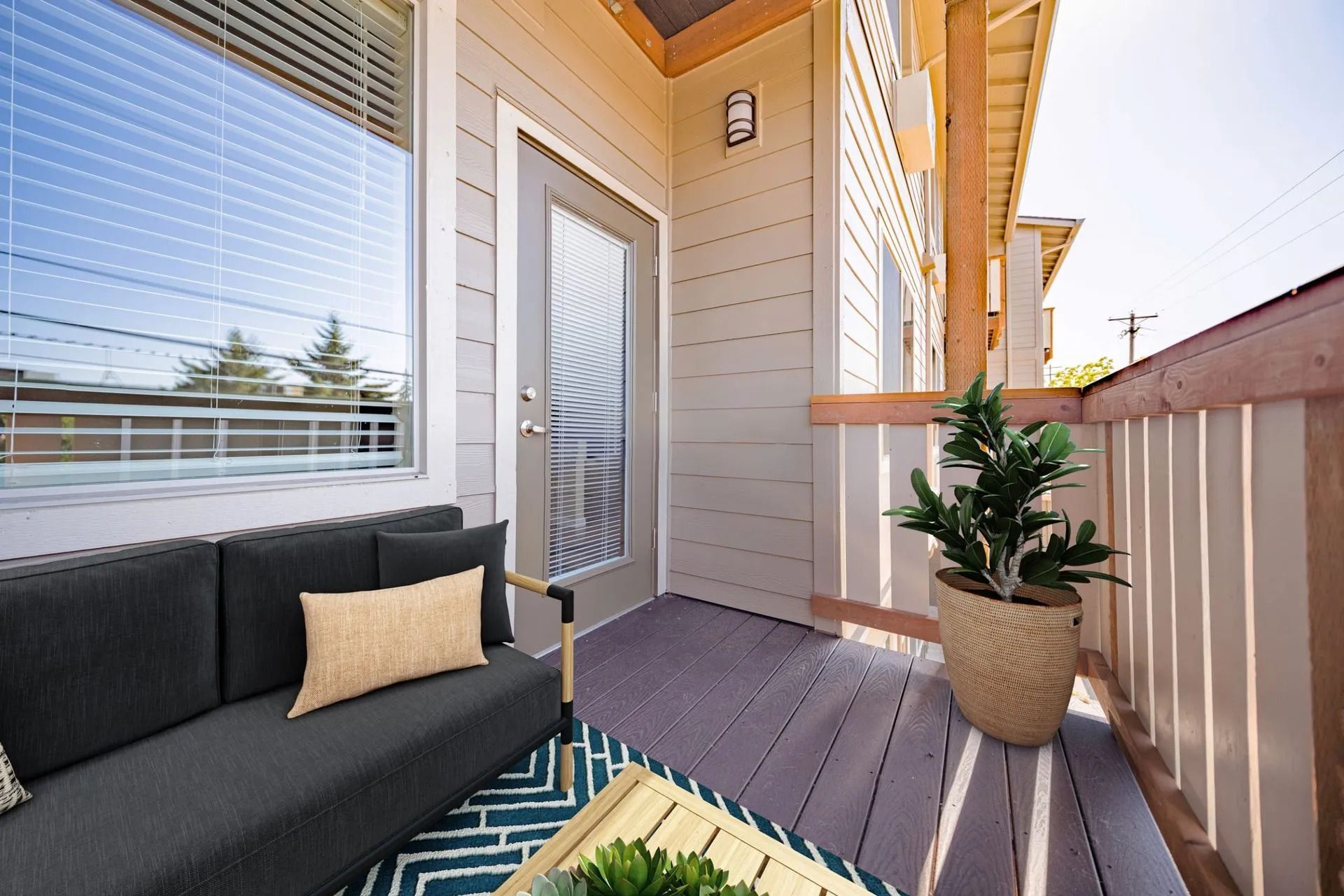 Modern apartment balcony with wicker sofa, potted plant, and striped rug in bright sunlight