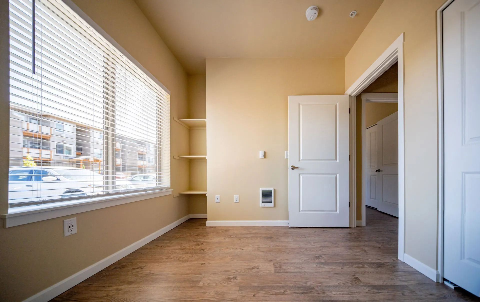 Empty beige bedroom with large window, wood floors, closet, and open bathroom door