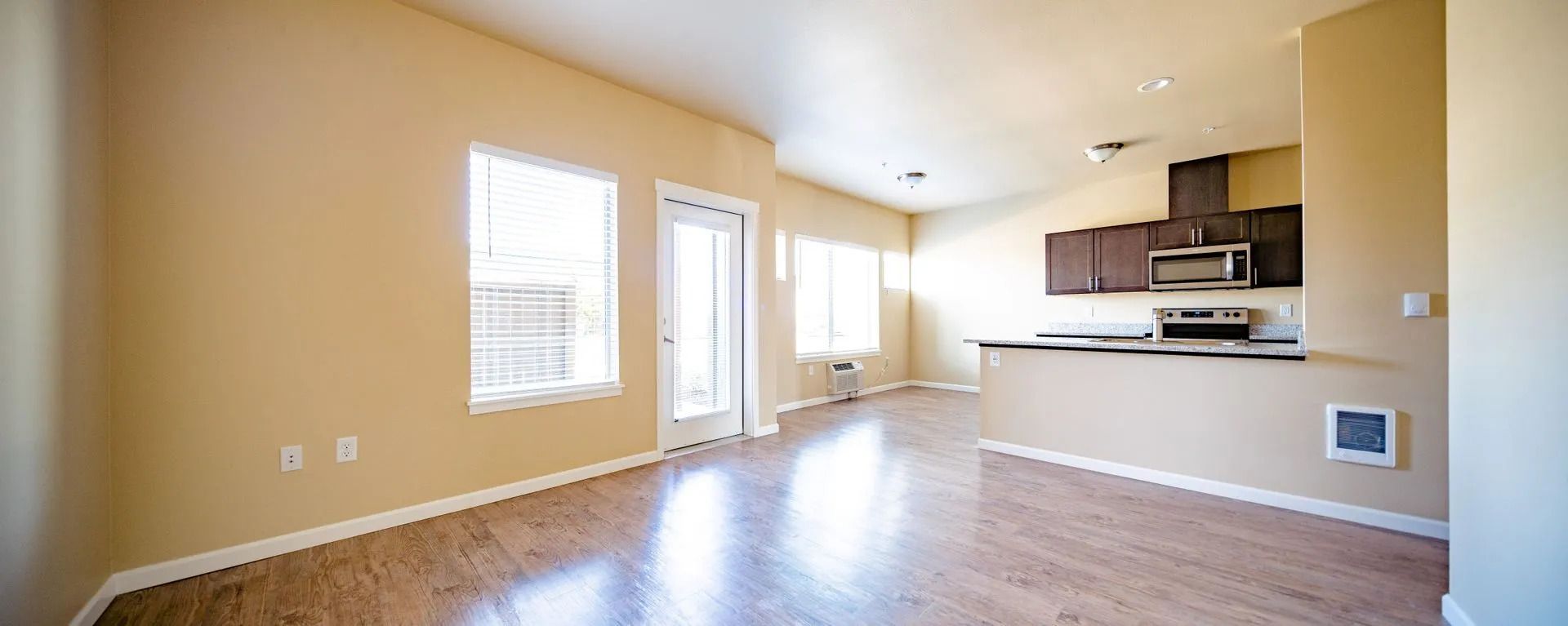 Empty unfurnished living room with tan walls, wood floors, and a kitchen area in the back