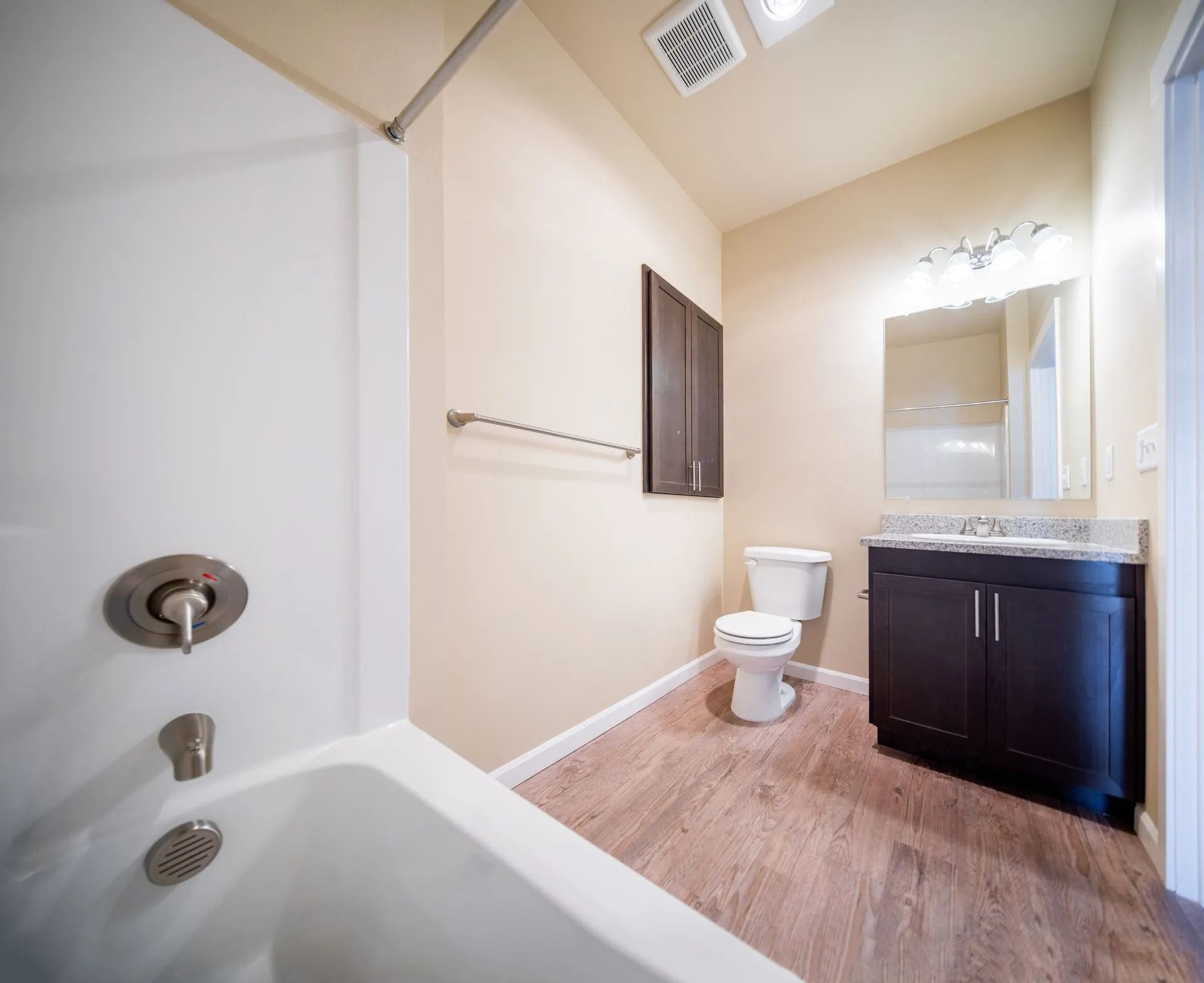 Small bathroom with tub, toilet, vanity sink, and mirror, in beige and dark wood tones.