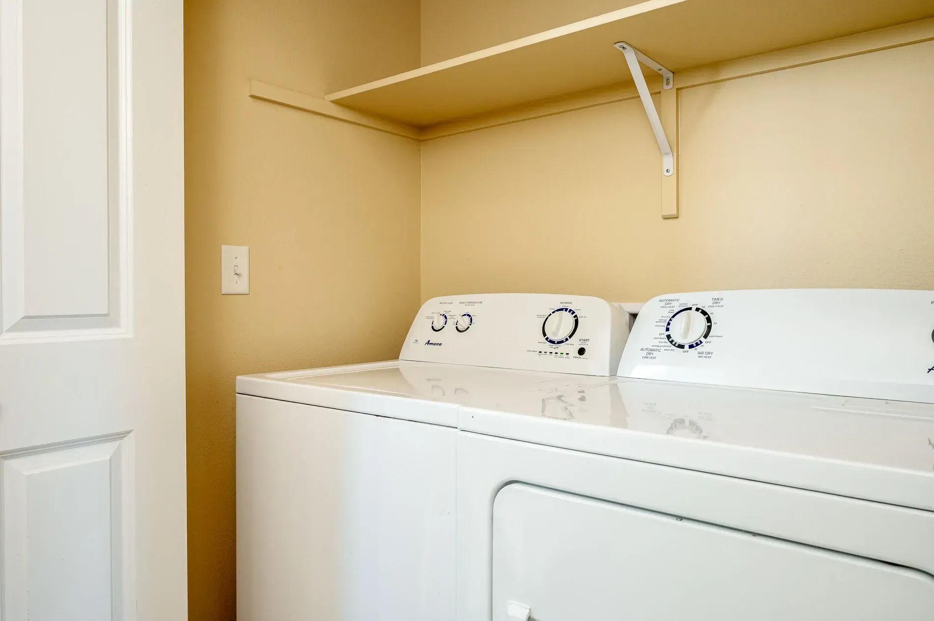 White washer and dryer in a beige laundry closet with a shelf overhead
