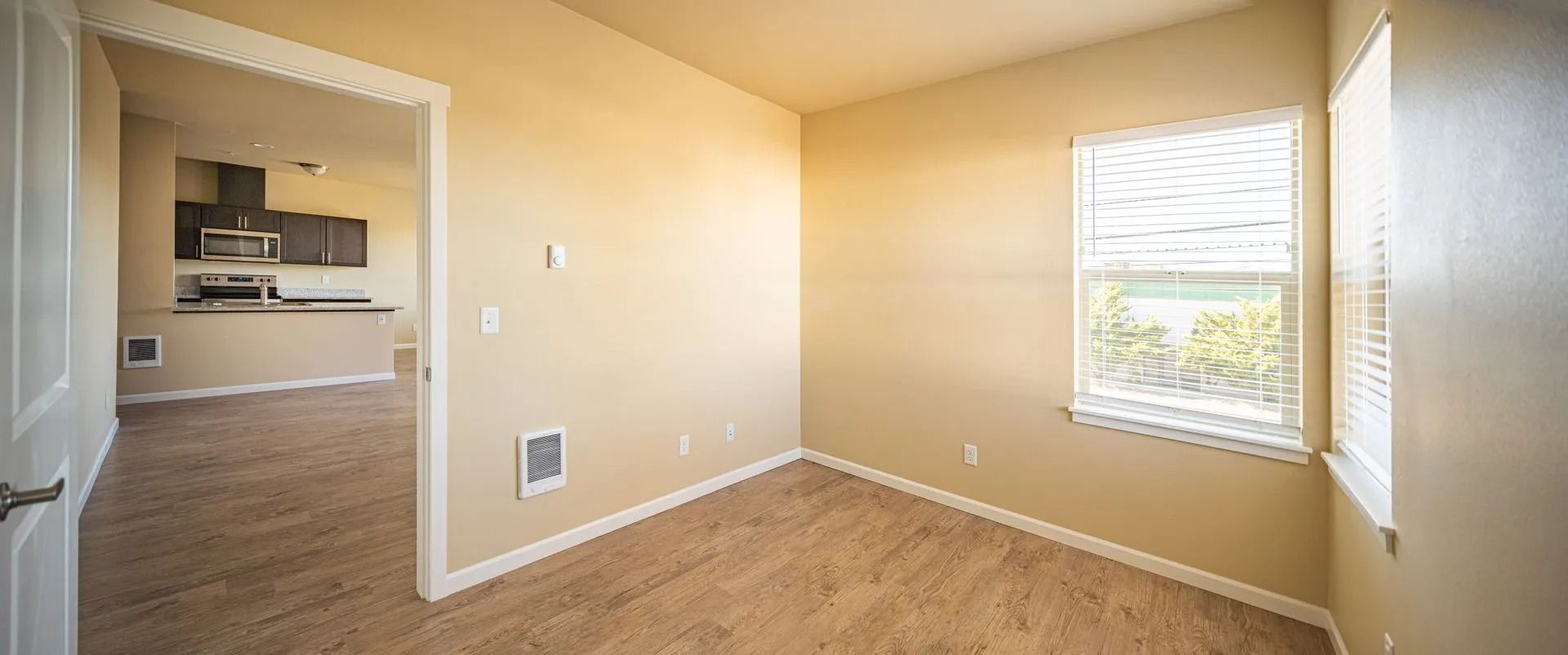 Empty beige room with carpet, window blinds, and open doorway to a kitchen area