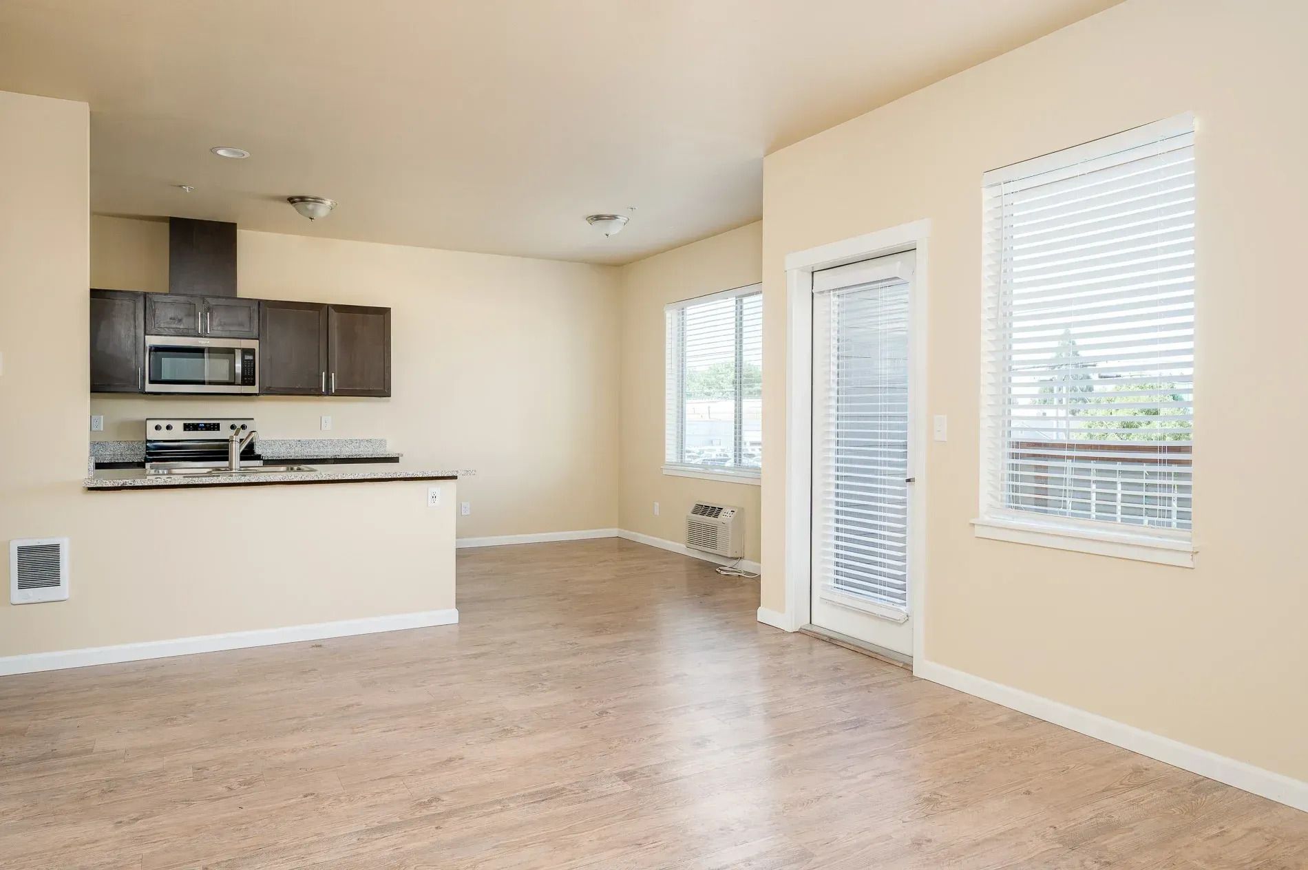 Empty beige living room with open kitchen, wood floor, and large windows with blinds