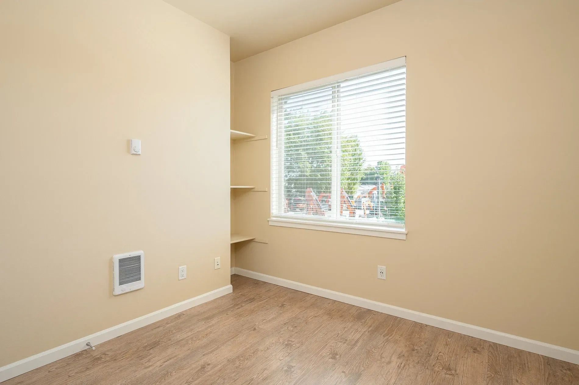 Empty beige bedroom with carpet, a window with blinds, and built-in corner shelves.