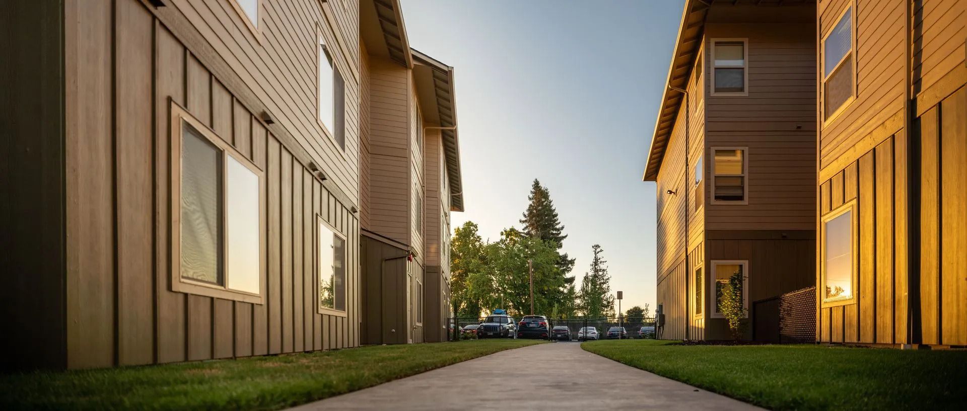 Sunlit apartment walkway between tan wooden buildings with a path leading to trees in the distance