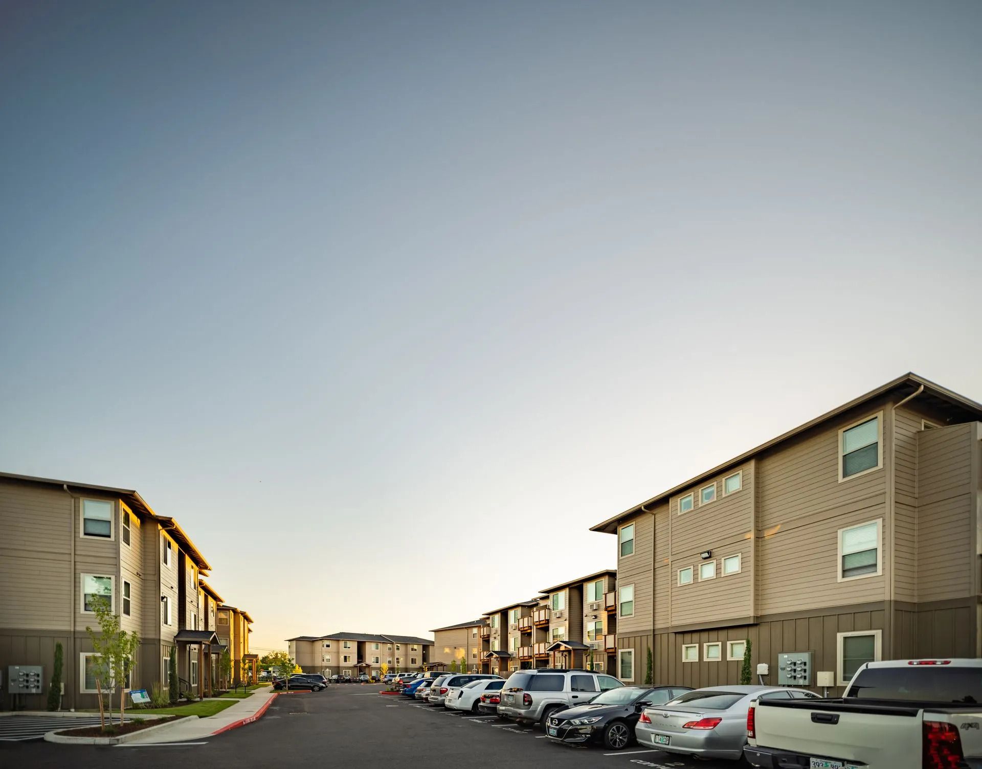 Apartment complex parking lot at dusk with parked cars and beige buildings lining a residential street