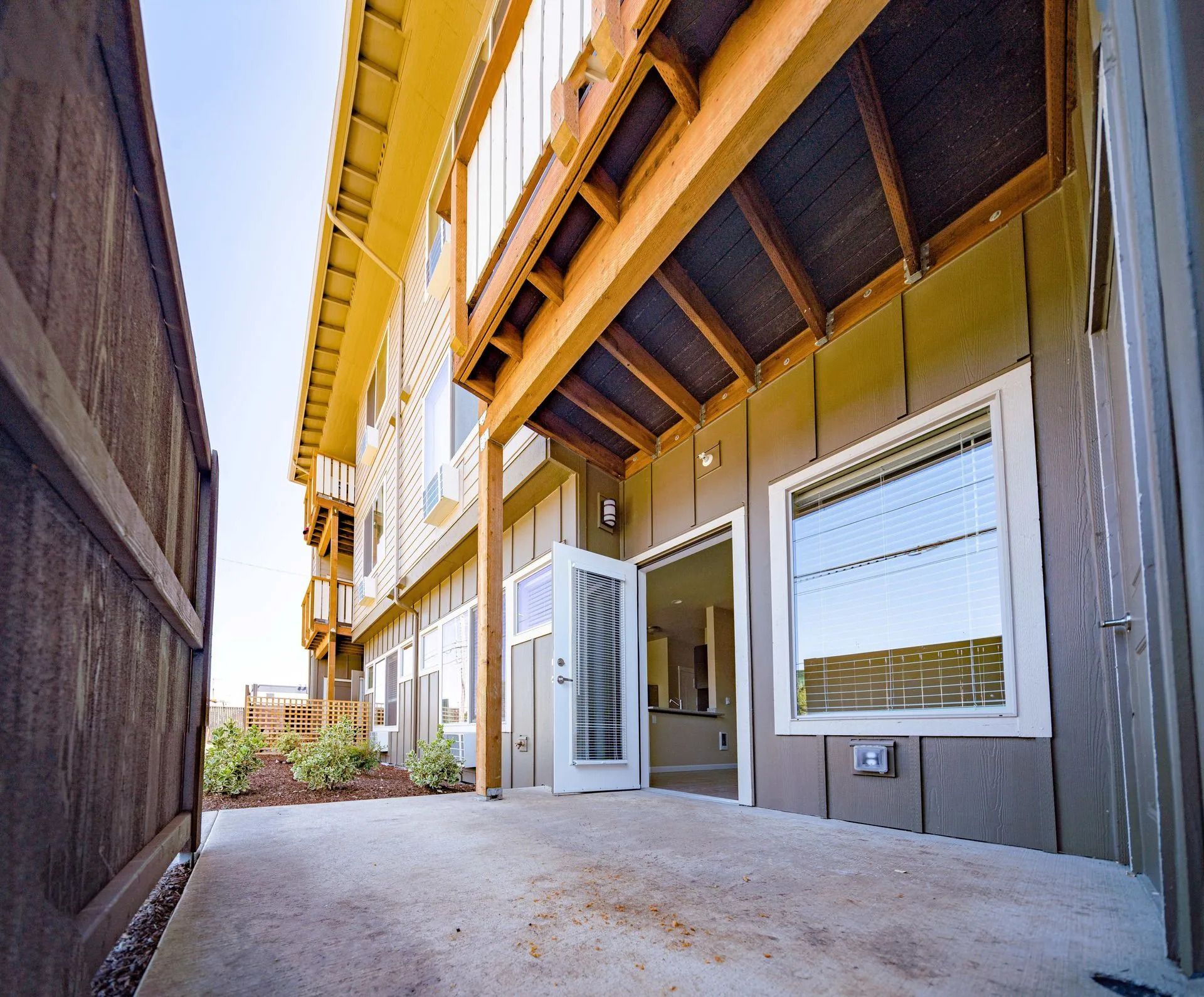 Narrow side yard beside a yellow house with an open white door and windows under an overhanging deck.