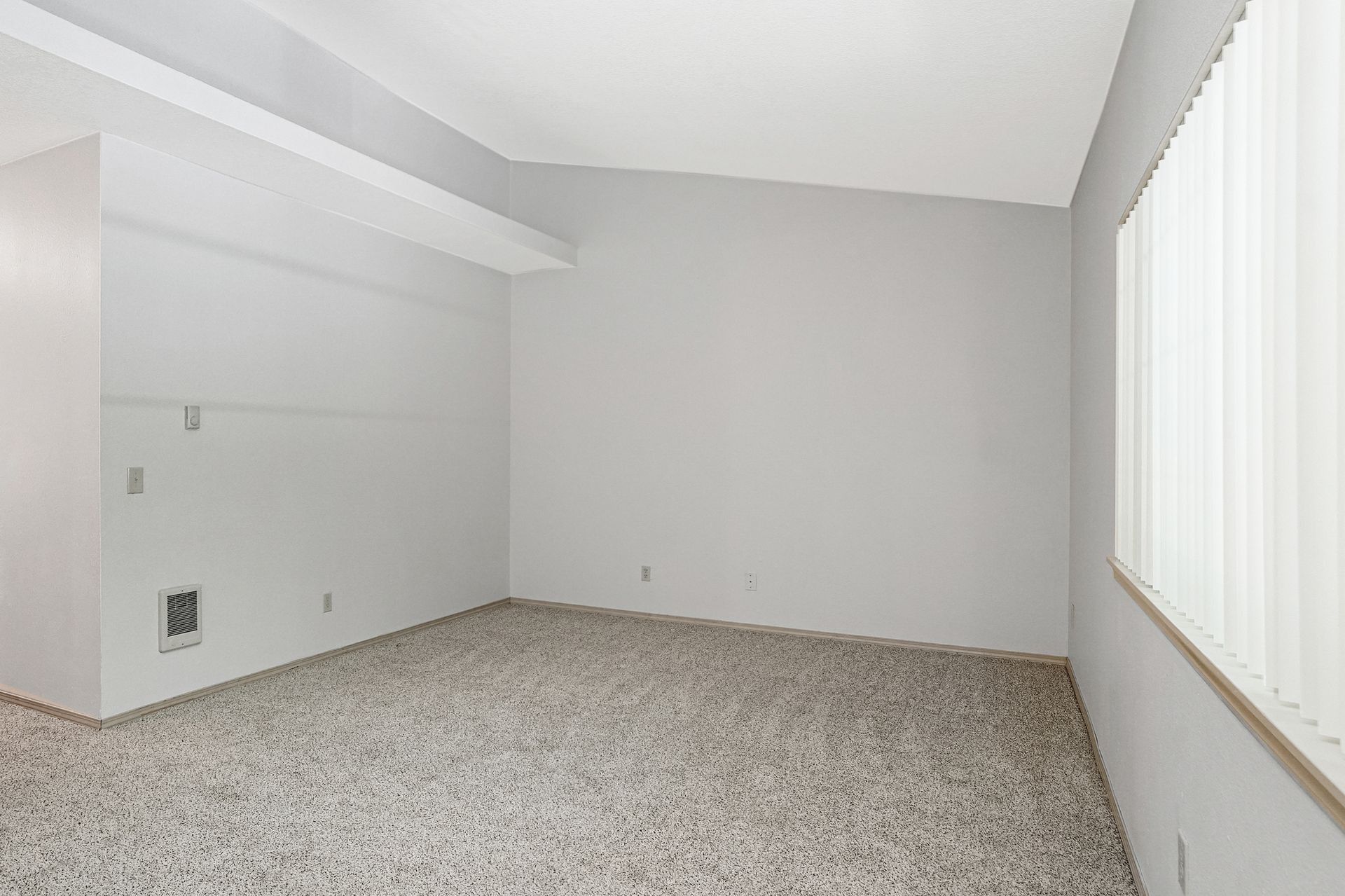Empty carpeted room with white walls and a large window with vertical blinds.