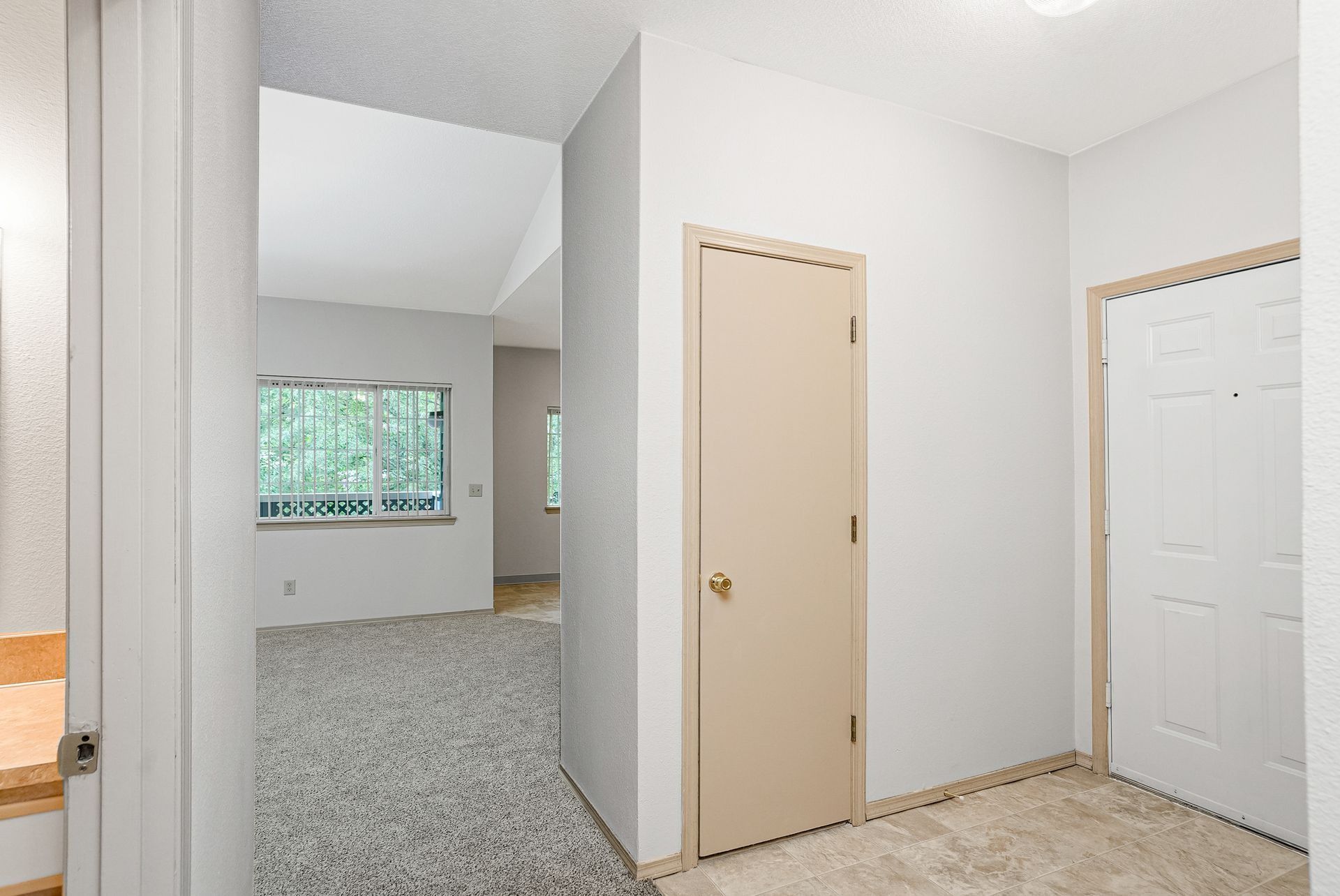 Empty beige hallway with white doors, carpeted room, and a window at the end