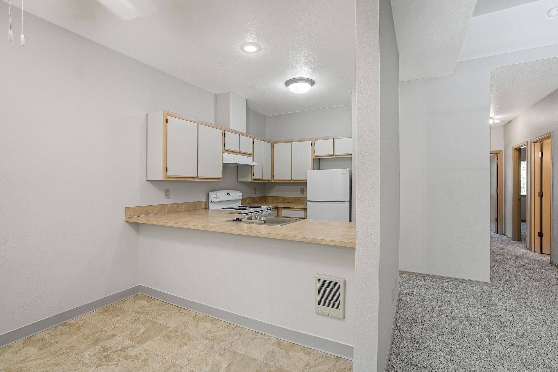 Small white kitchen with tan counters opening to a carpeted hallway in an empty apartment