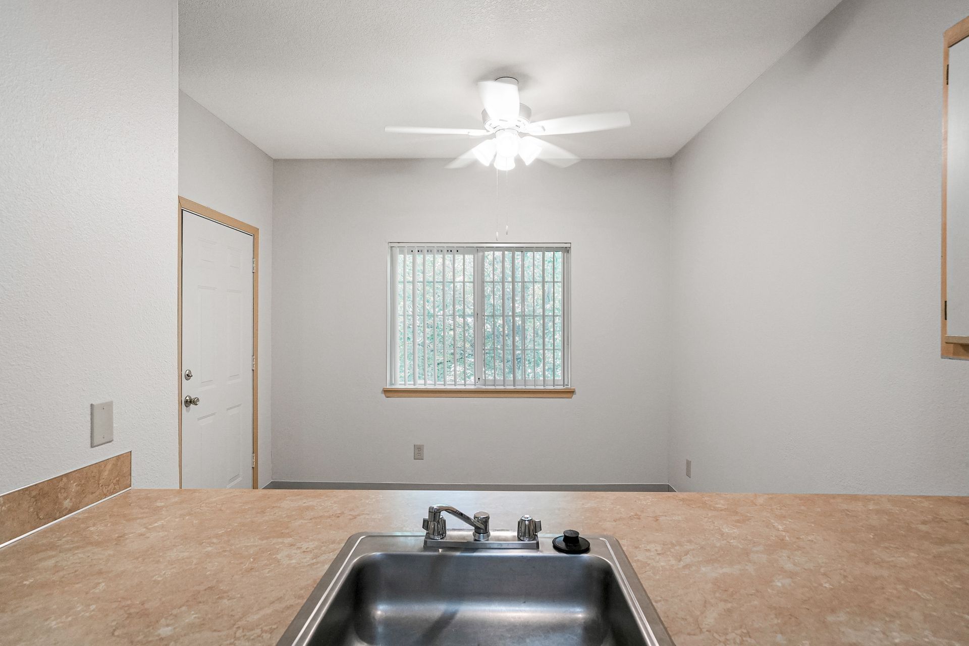 Empty kitchen with beige countertop, sink, white walls, ceiling fan, and window.