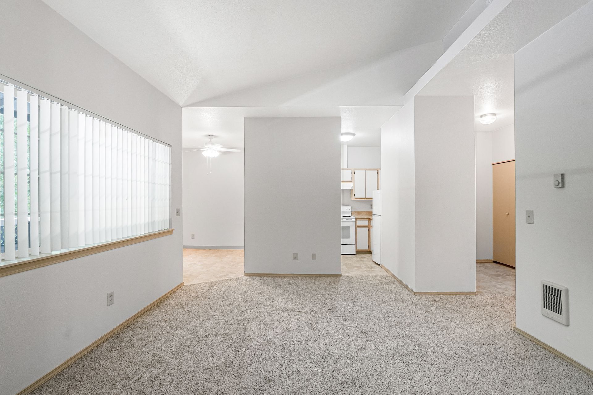 Empty carpeted living room with white walls, large window blinds, and a doorway to a kitchen area