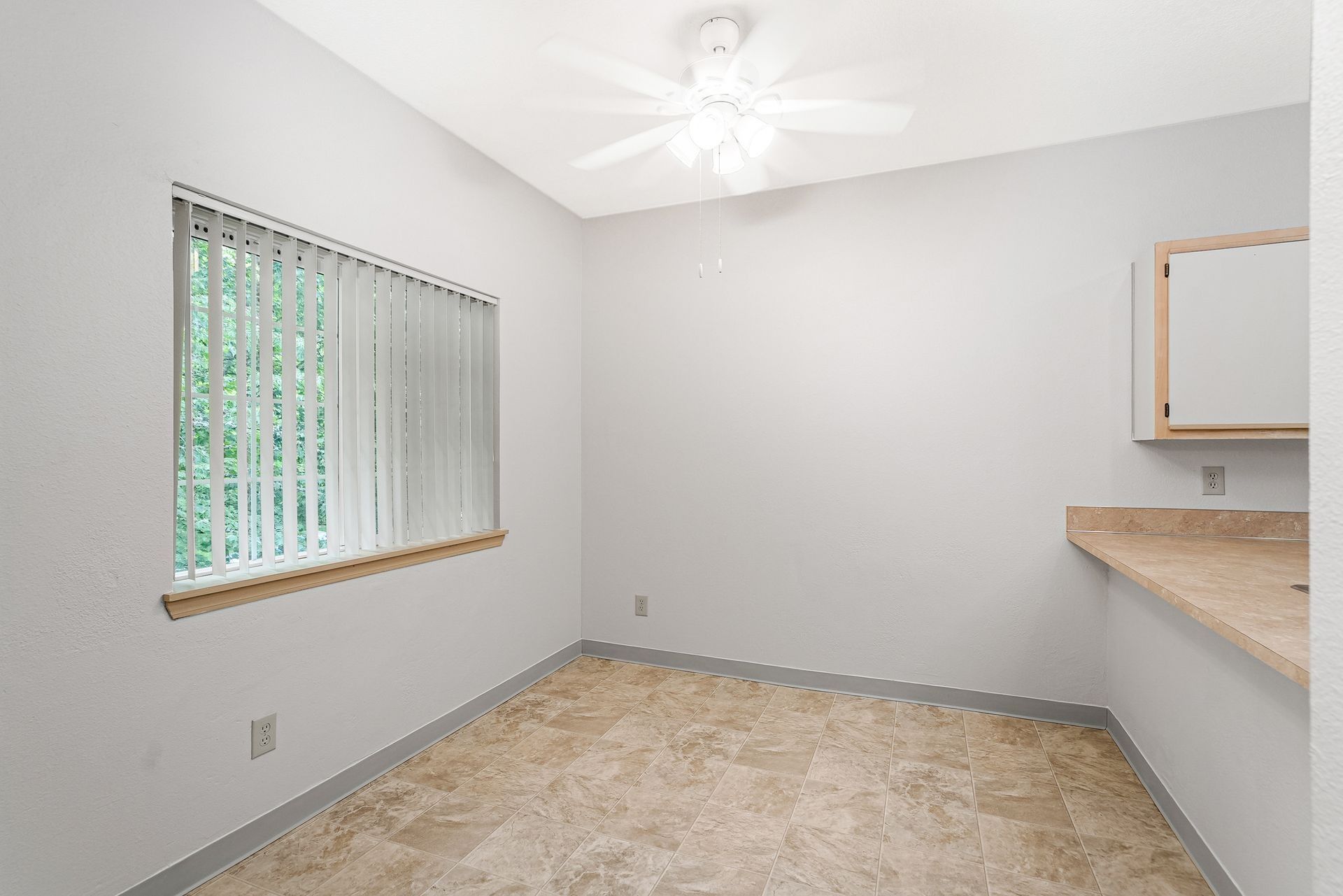 Empty beige room with window blinds, ceiling fan, and a small counter pass-through