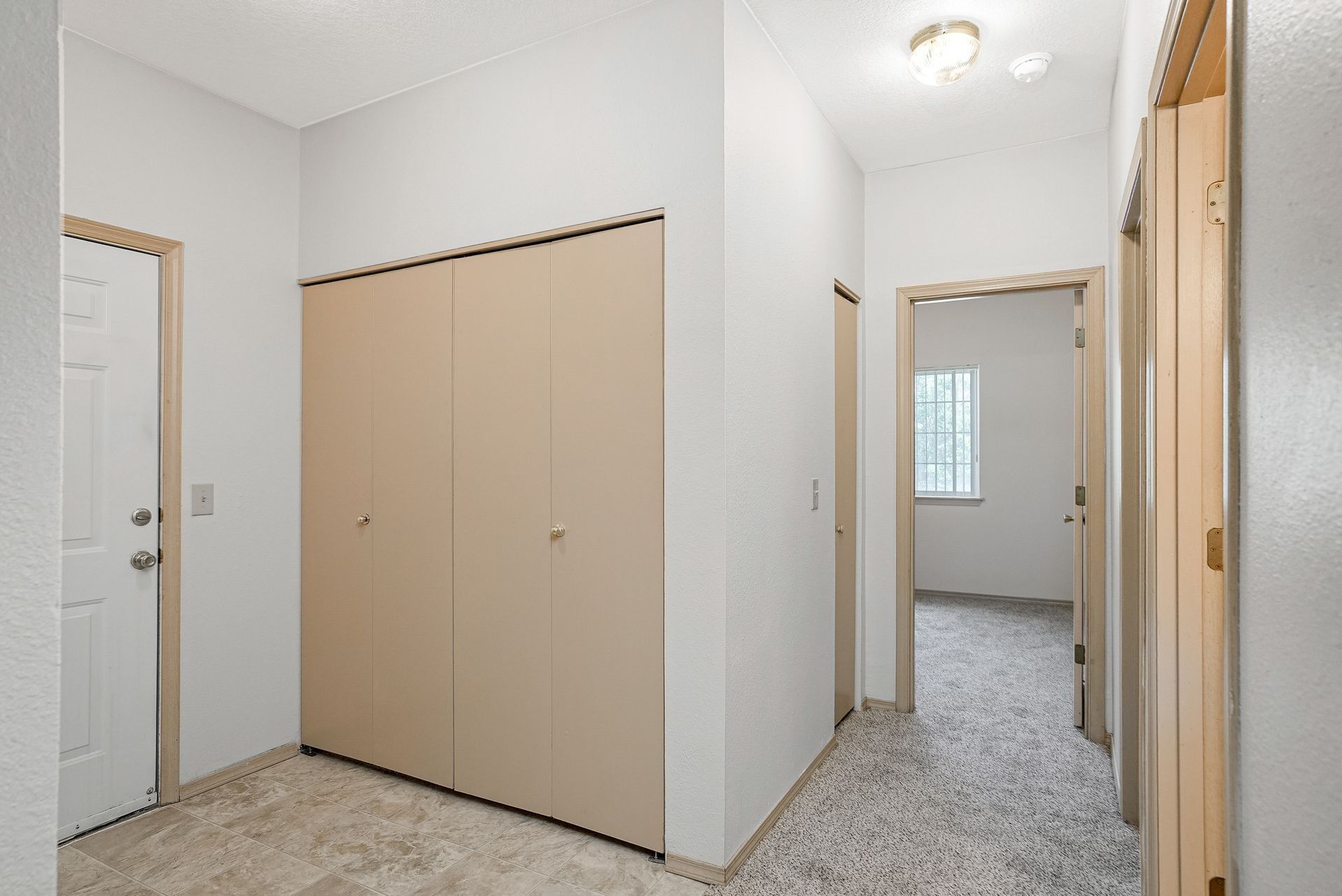 Empty hallway with beige closets, white walls, and carpeted floor leading to a room.