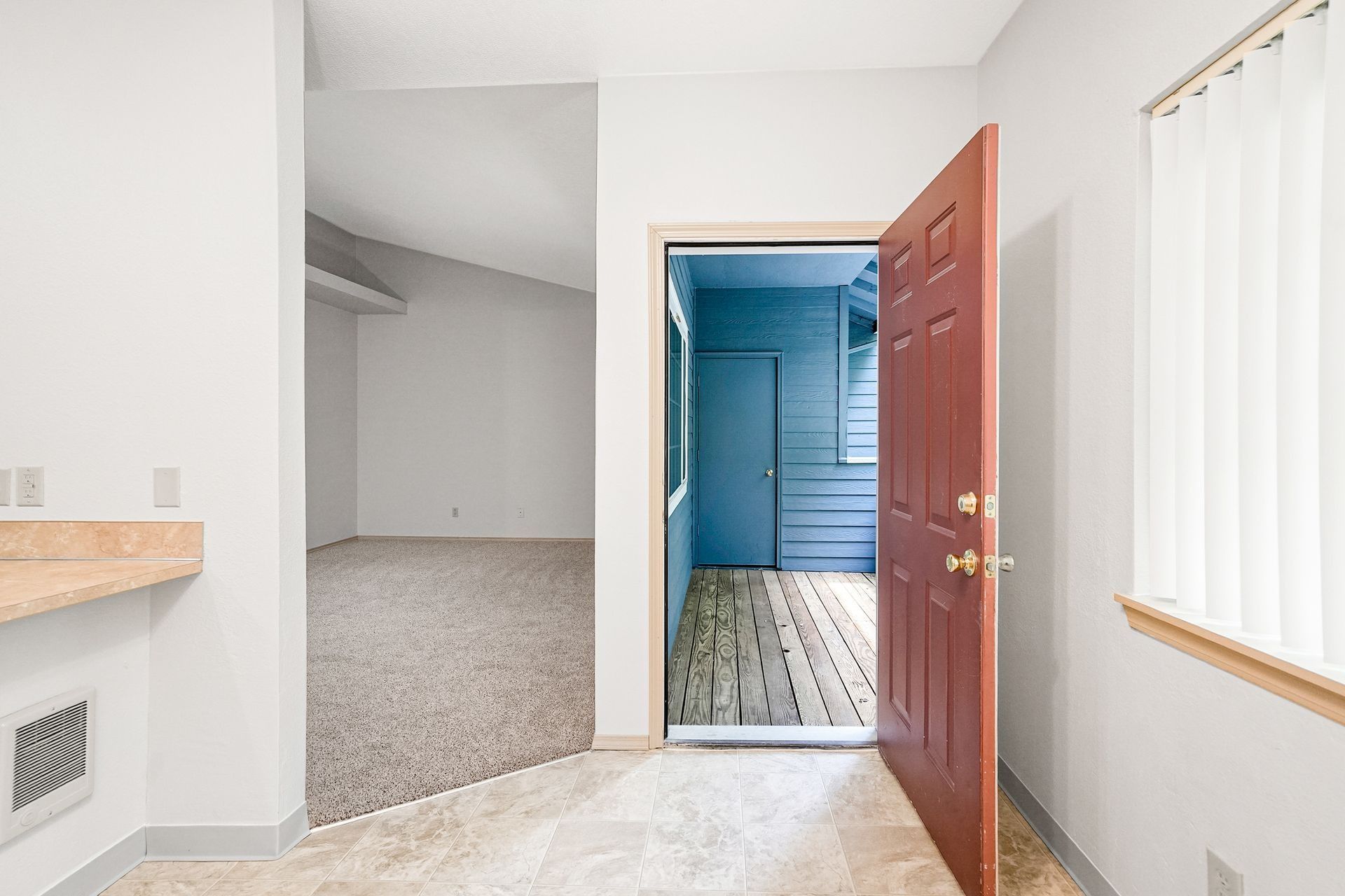 Bright entryway with open wooden door, tiled floor, carpeted room, and white blinds on the right.