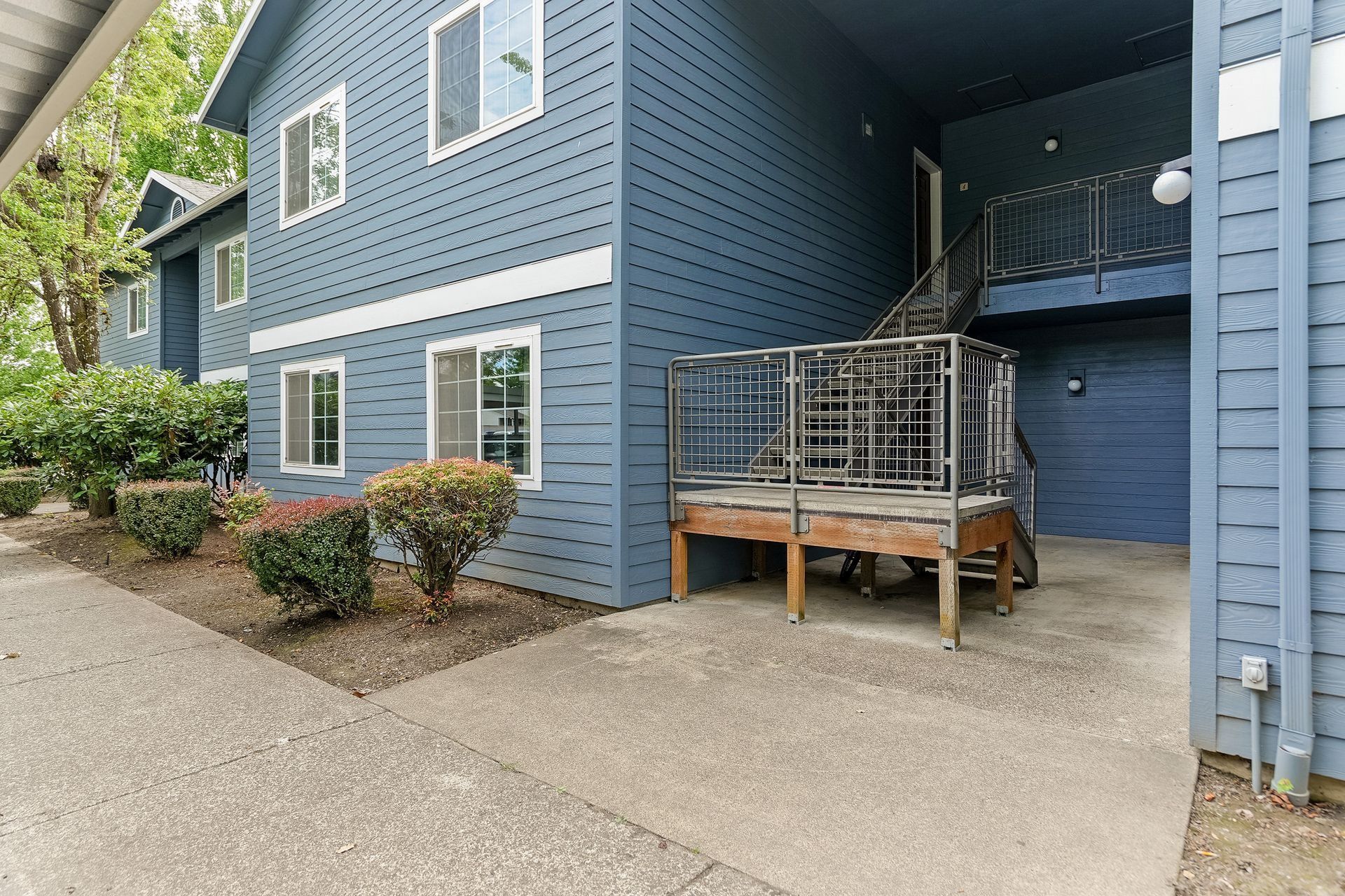 Blue apartment exterior with a small wooden deck and ramp beside a concrete walkway