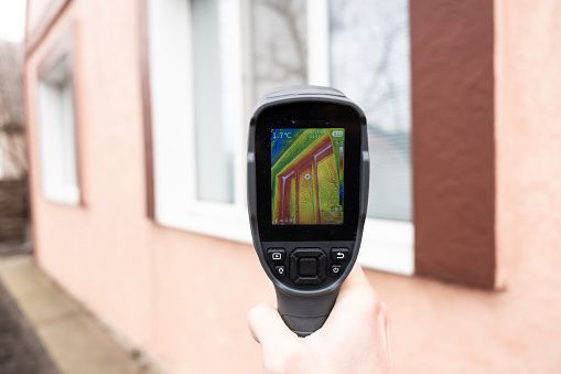 Handheld thermal camera aimed at a building wall, displaying a heat map on its screen