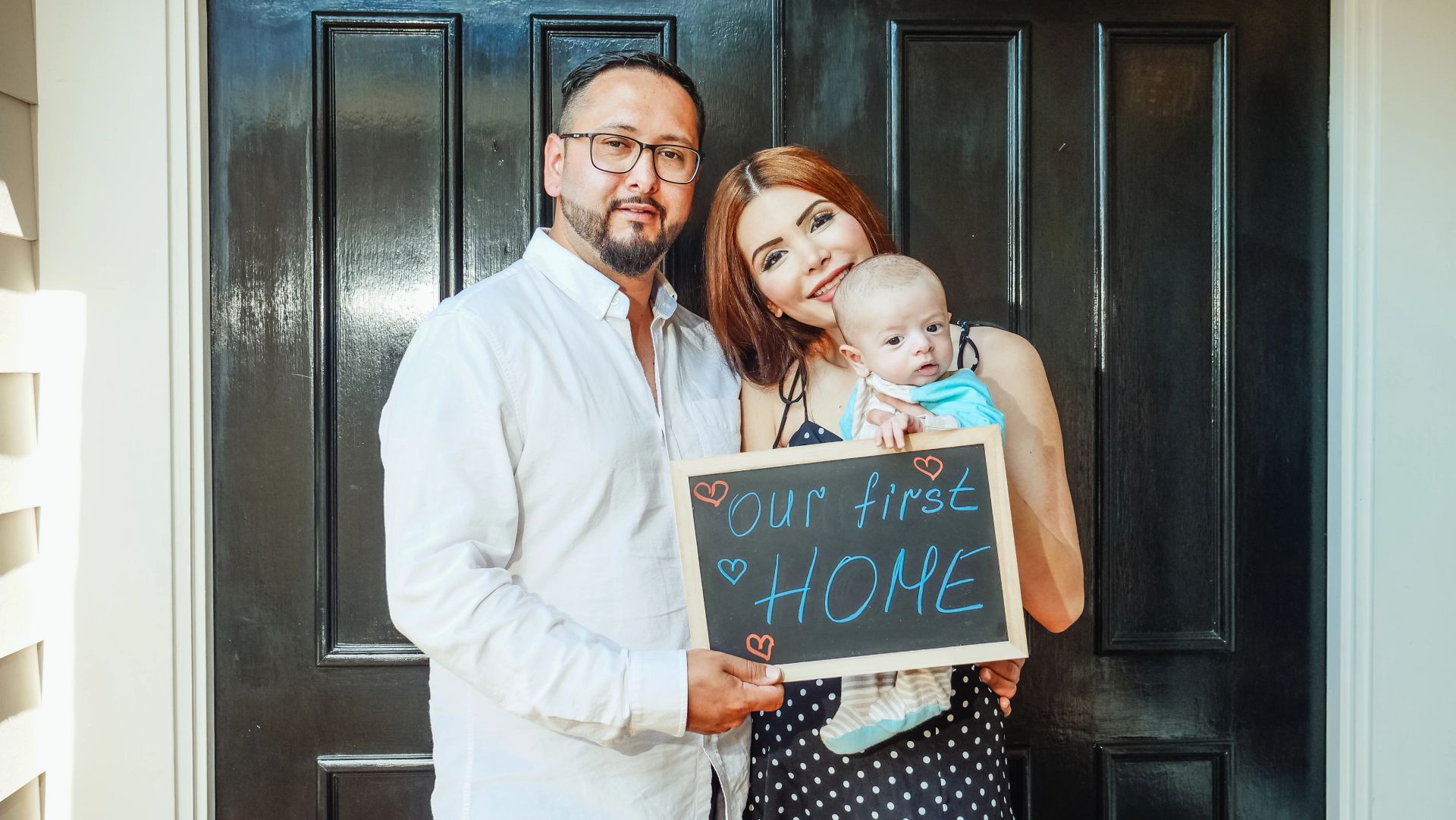Family holding a chalkboard sign that reads “Our New Home” in front of a black front door
