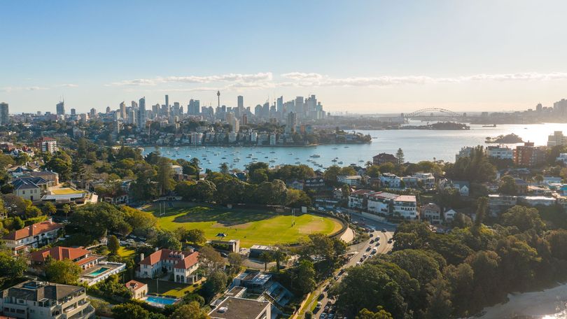 Aerial view of a coastal park with city skyline, harbor, and sunlight over trees and water