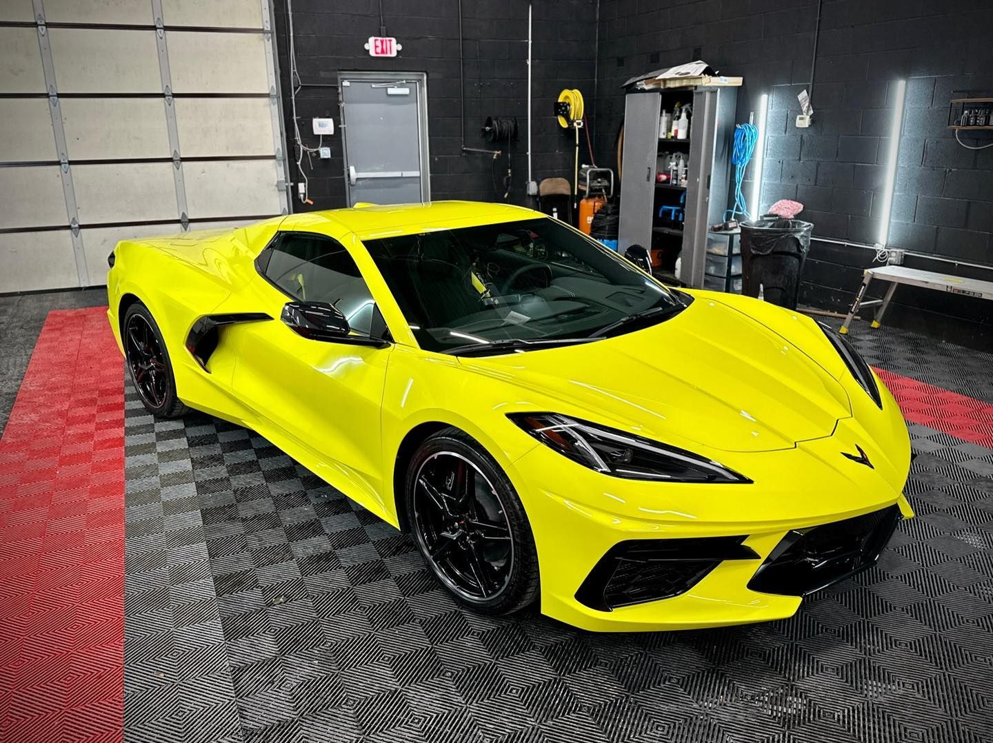 Bright yellow sports car in a garage, angled front view on a black patterned floor.