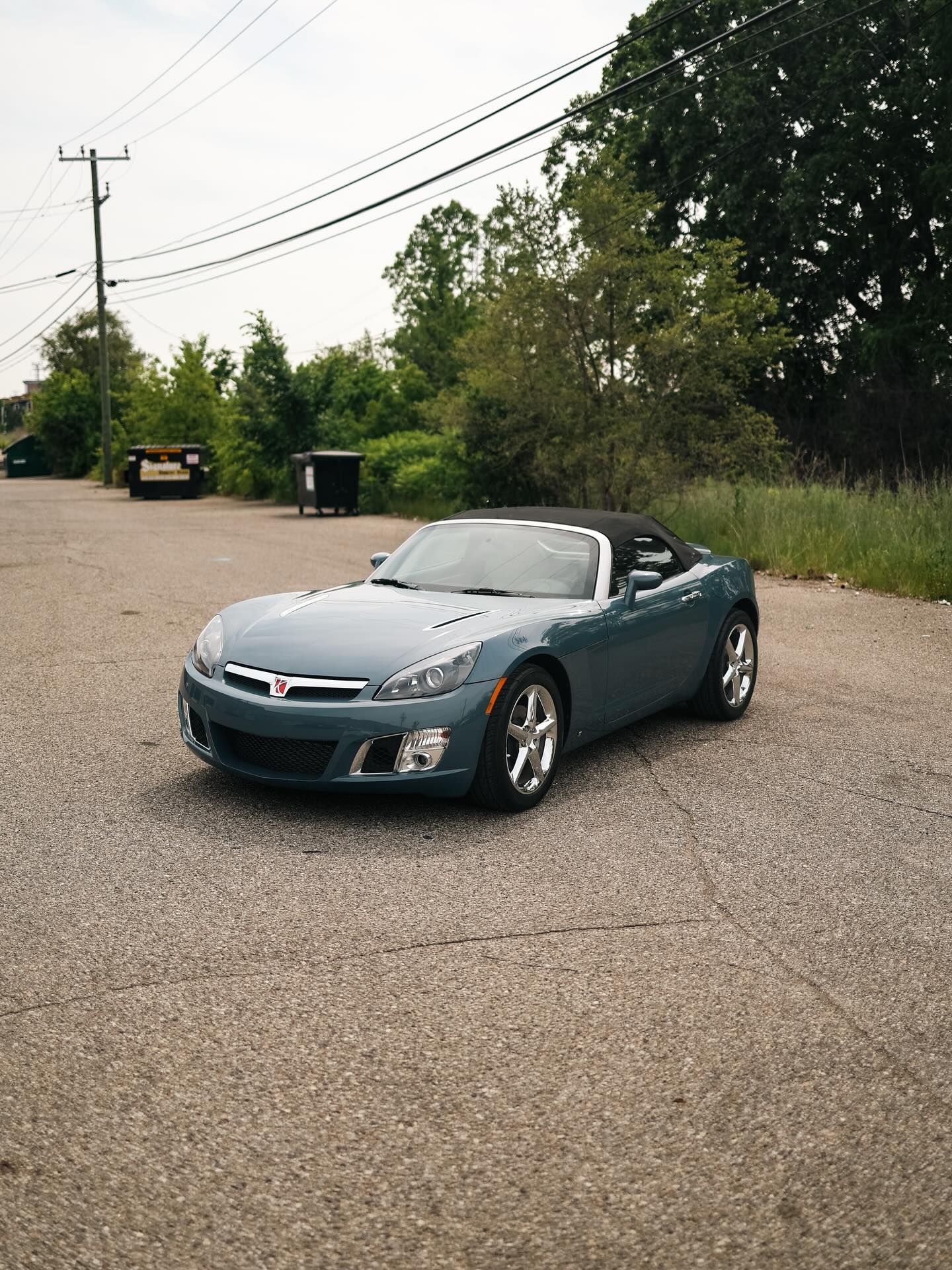 Blue sports car parked on a gravel lot beside trees and power lines