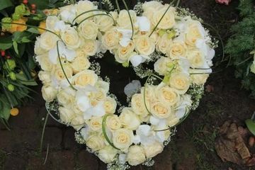 A heart-shaped funeral wreath made of cream-colored roses and white orchids arranged on a dark brick surface.