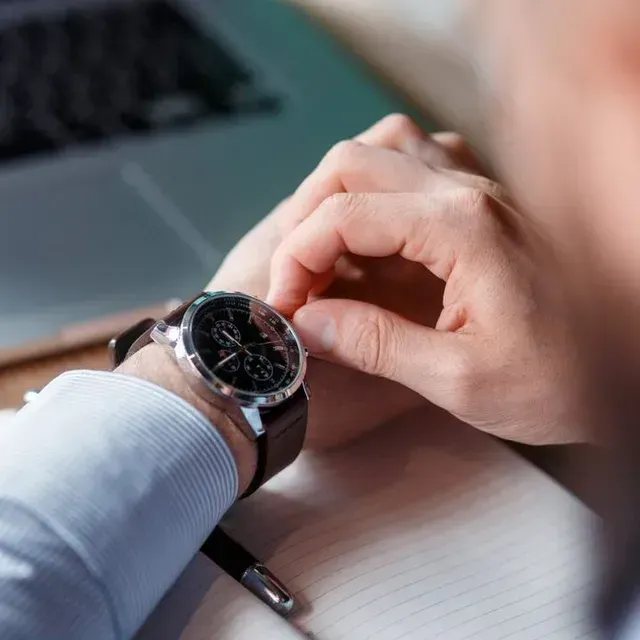 A person in a blue shirt adjusting a black chronograph watch on their wrist while seated at a desk with a laptop.