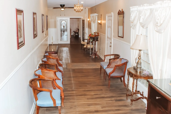 Long, narrow hallway with wooden chairs, framed art, tables, and white curtains along the walls
