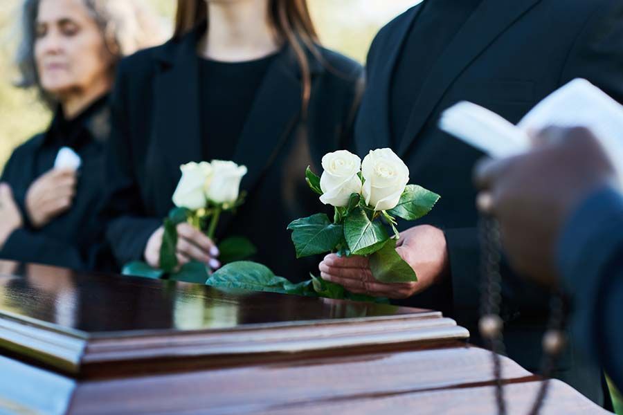 Mourners in dark attire hold white roses over a casket at an outdoor funeral service.