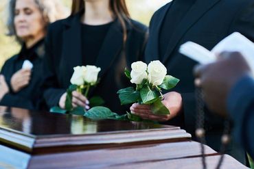 Mourners in dark attire hold white roses over a casket at an outdoor funeral service.