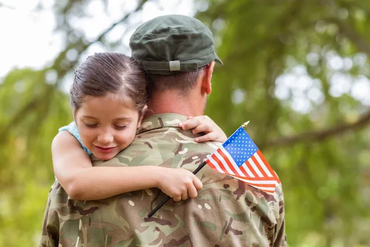 A person in a military uniform embraces a child who is holding a small American flag in an outdoor, wooded setting.
