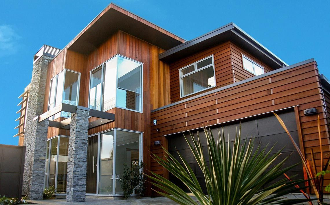 Modern two-story house with wood siding, stone columns, and large windows under a blue sky.