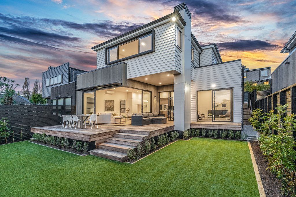 Modern two-story house with wooden deck, steps, and green lawn, under a sunset sky.