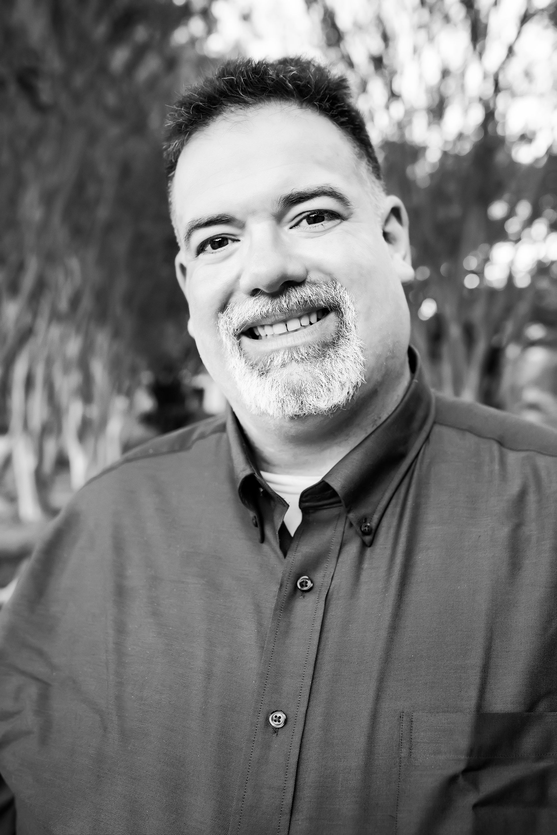 Man with short, dark hair and salt-and-pepper beard smiles broadly while wearing a button-down shirt outdoors.