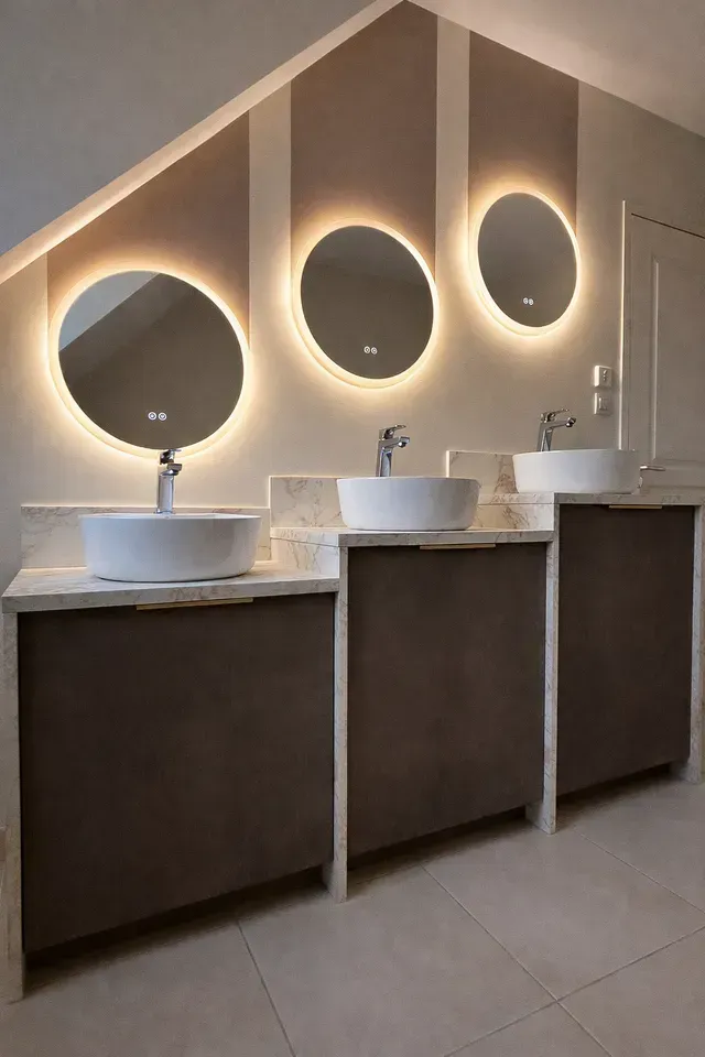 Toilettes modernes avec trois miroirs ronds rétroéclairés au-dessus de lavabos blancs et de meubles-lavabos en bois foncé sous un plafond incliné