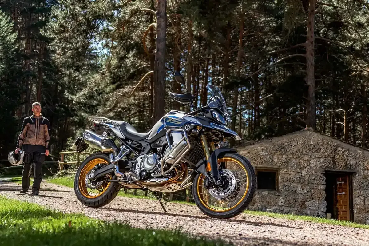 Motocicleta azul e dourada estacionada em uma trilha na floresta ao lado de uma pessoa e uma cabana de pedra.