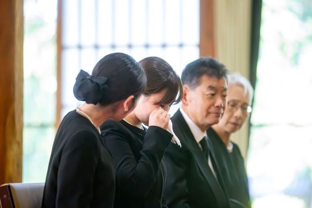 Four people in black attire sit in a row indoors, one dabbing tears with a tissue.